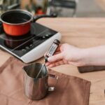 A person measures the temperature of melted wax on an electric stove, highlighting modern craft techniques.