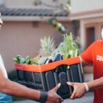 Two men exchanging a crate of fresh vegetables during a contactless delivery outdoors with safety measures.