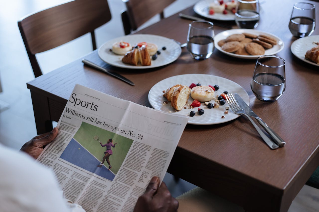 services-04 A man reads the sports section while enjoying breakfast at a neatly set table indoors.
